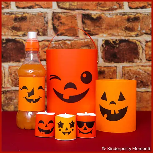 Orangefarbene Halloween-Dekorationen mit schwarzen lachenden Gesichtern, darunter eine Flasche, eine Laterne, ein Windlicht und drei kleine Teelichtkerzen vor Ziegelsteinwand