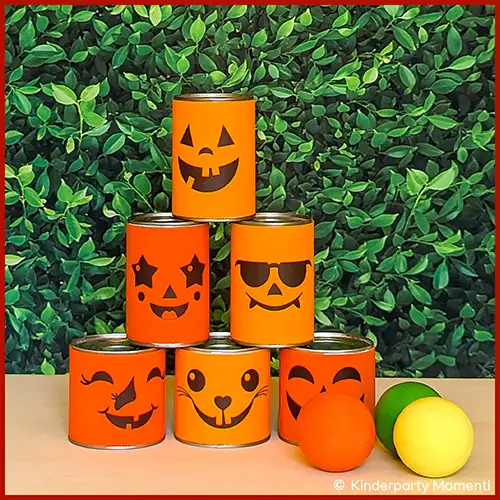 Sechs orangefarbene Dosen mit verschiedenen Halloween-Gesichtern als Dosenwerfen Spiel vor grünem Hintergrund und drei bunten Bällen