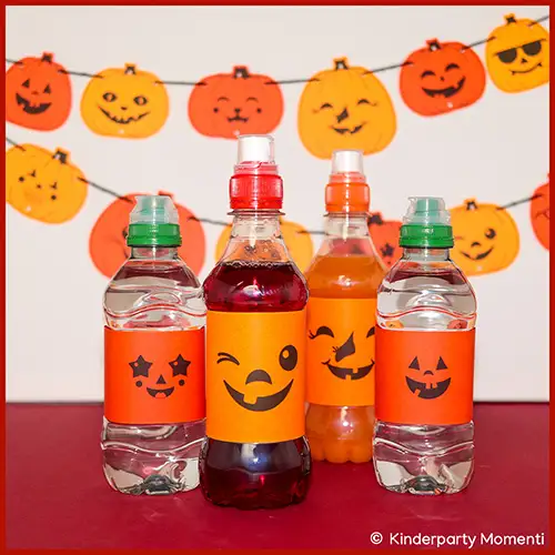 Vier kleine Flaschen mit orangefarbenen Halloween Flaschenetiketten, darauf lachende Kürbisgesichter, im Hintergrund Kürbisgirlanden an der Wand