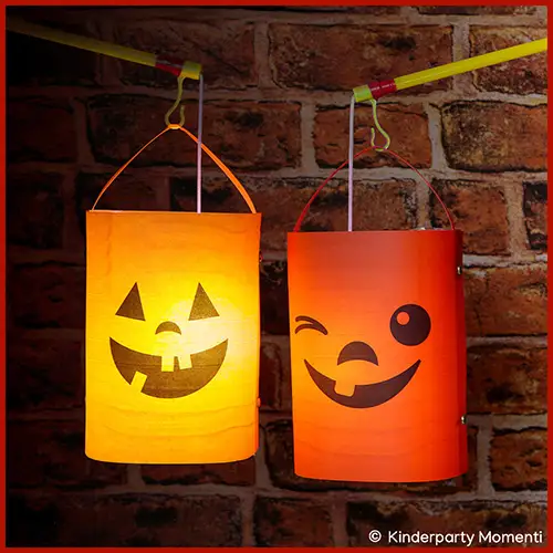 Zwei orangefarbene, selbstgebastelte Halloween Laternen mit lachenden Kürbisgesichtern hängen vor einer Ziegelsteinwand.