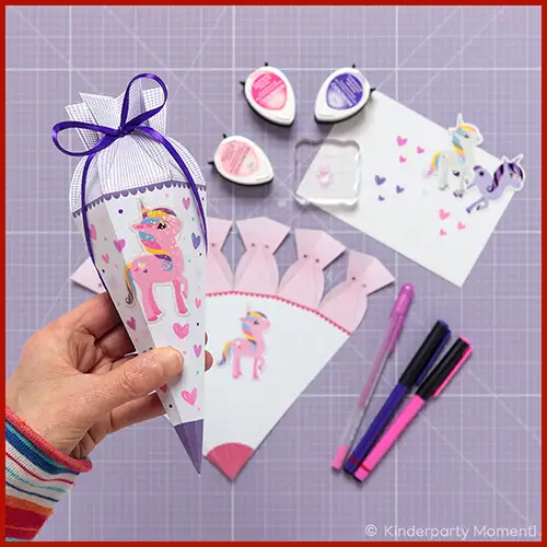 Eine Hand hält eine kleine selbstgebastelte Lila Schultüte, auf der ein Einhorn Aufkleber zu sehen ist. Im Hintergrund ist die Schultüten Vorlage und Bastelwerkzeug zu sehen.