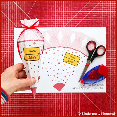 Eine Hand hält eine kleine Schultüte mit einem Ortsschild. Im Hintergrund liegen Bastelmaterial und eine Schultüten Vorlage