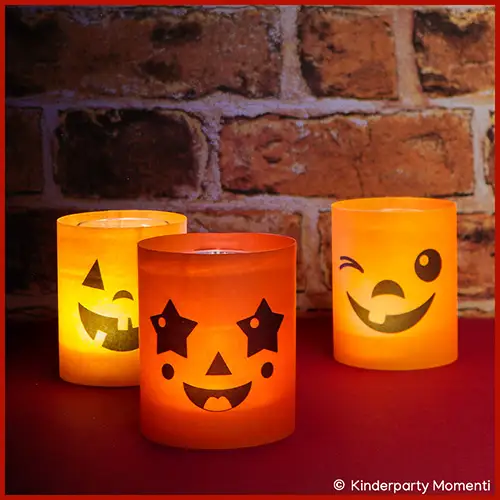 Drei leuchtende Windlichter aus Transparentpapier mit lachenden Halloween-Gesichtern vor einer Backsteinwand