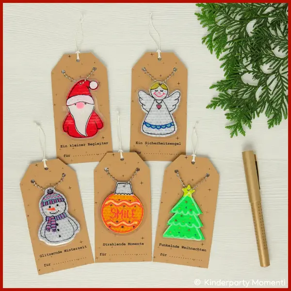 Fünf reflektierende Weihnachtsanhänger mit Motiven: Weihnachtsmann, Engel, Schneemann, Weihnachtskugel mit 'SMILE' und Weihnachtsbaum, alle mit Kugelketten an Geschenkanhängern befestigt.