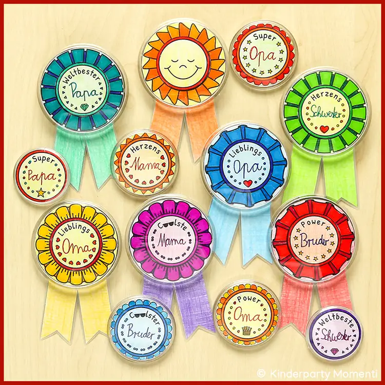 Kleines DIY Geschenk - Orden Button selber machen ohne Maschine Bunte selbstgemachte Anstecker Button mit handgeschriebenen Texten wie 'Super Papa', 'Lieblings Oma', 'Herzens Mama' und 'Power Bruder' auf hellem Untergrund