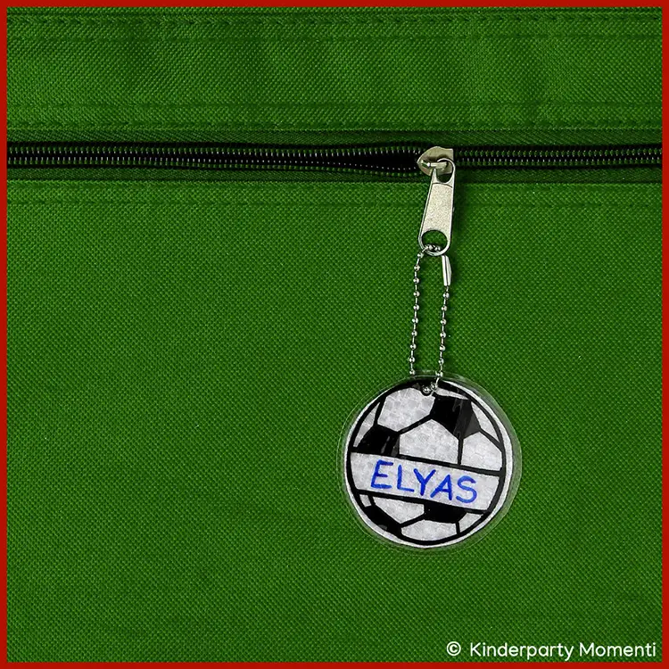 Personalisierter Fußball Anhänger als DIY Reflektor Anhänger in Form eines Fußballschuhs mit schwarzen Nocken und roten sowie blauen Streifen an einem grünen Rucksack