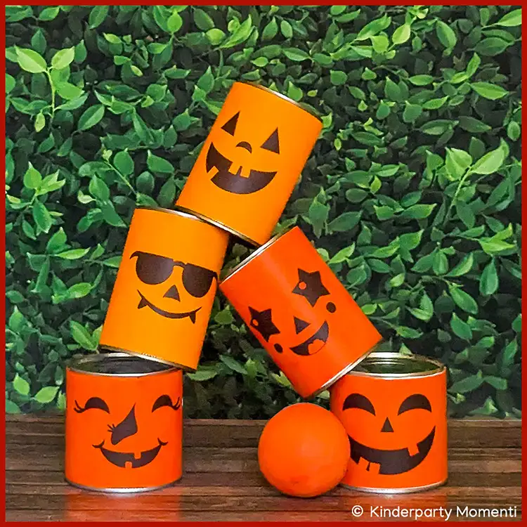Dosenwerfen Spielen bei Kinderfesten Fünf Dosen mit verschiedenen lachenden Halloween-Gesichtern in Orange zu einem Pyramidenstapel auf Holz vor grünem Hintergrund und einem orangenen Ball davor