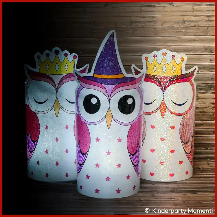 Drei leuchtende Eulen DIY Windlichter aus Papier, zwei mit goldener Krone und eine mit lila Hexenhut, vor Holzwand.