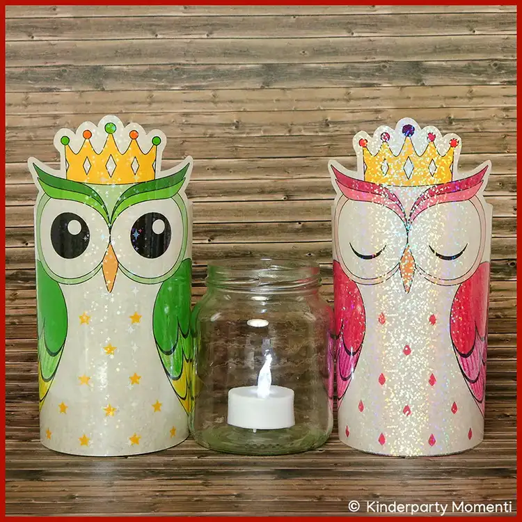 Zwei glitzernde DIY Eulen Windlichter mit Kronen, eine grün und eine rosa, flankieren ein Glas mit einer brennenden Teelichtkerze vor Holzwand.