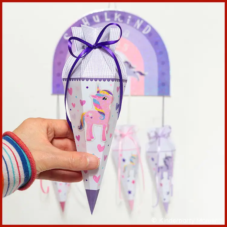Kleine Lila Schultüte mit Einhorn Lila Schultüte, auf der ein Einhorn Aufkleber zu sehen ist. Im Hintergrund hängt ein Regenbogen, an dem kleine Papierschultüten hängen.