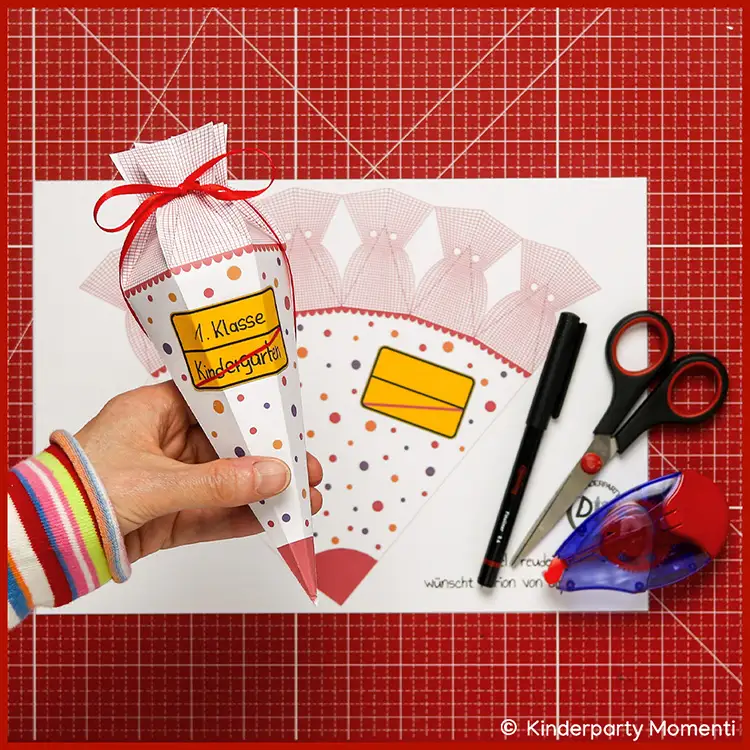 Kleine Schultüte basteln mit Ortsschilder zum selber beschriften Hand hält eine gebastelte kleine Schultüte aus Papier mit Aufschrift '1. Klasse Kindergarten' vor einem Bastelbogen, daneben liegen Schere, Stift und Kleberoller auf roter Bastelmatte.