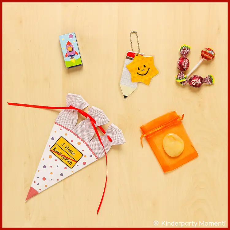 Ideen zum Füllen für Minischultüte Kleine gebastelte Schultüte mit rotem Band und Aufschrift '1. Klasse Kindergarten' neben einem Reflektor in Form eines Bleistifts mit gelbem Stern und einem orangefarbenen Organzasäckchen mit einem Stein sowie Süßigkeiten.