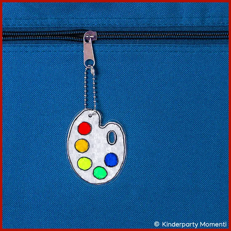 Farben Mischpalette als reflektierender Taschenanhänger Reflektor Anhänger in Form einer Malkünstlerpalette mit bunten Farbflecken an einem Reißverschluss einer blauen Tasche