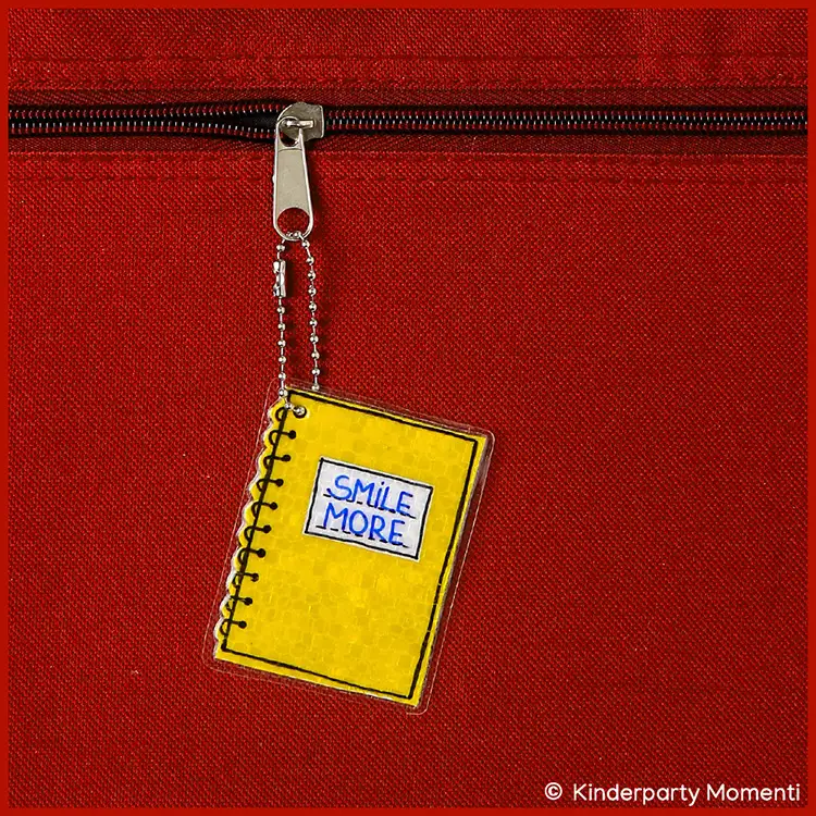 Kleines Notizbuch als DIY Reflektor Gelber Notizbuchanhänger mit der Aufschrift 'SMILE MORE' an einer Kugelkette an einem Reißverschluss einer roten Tasche