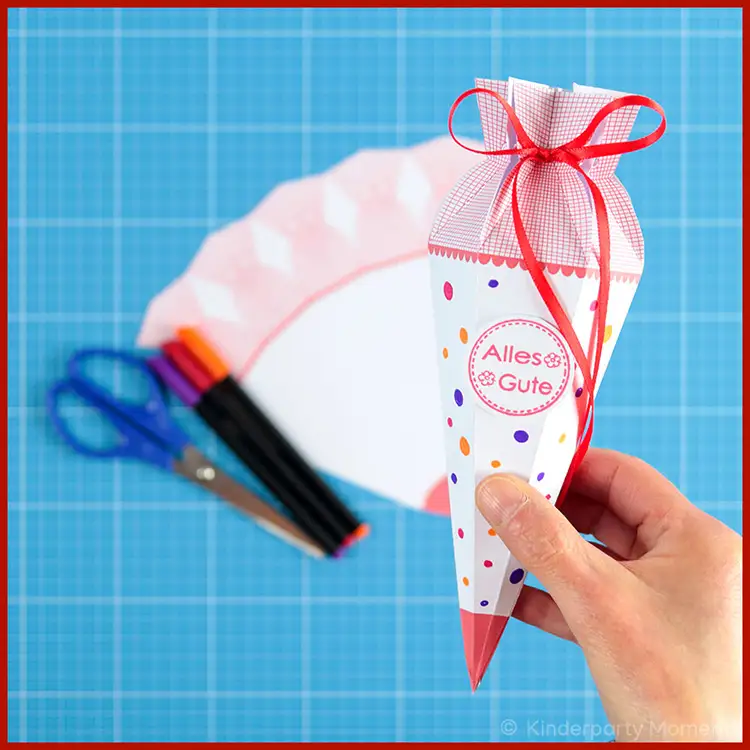 Kleine Papier Schultüte selber machen - 6 eckig Eine Hand hält eine kleine selbstgebastelte Schultüte auf der 'Alles Gute' steht. Im Hintergrund ist die Schultüten Vorlage und Bastelwerkzeug zu sehen.
