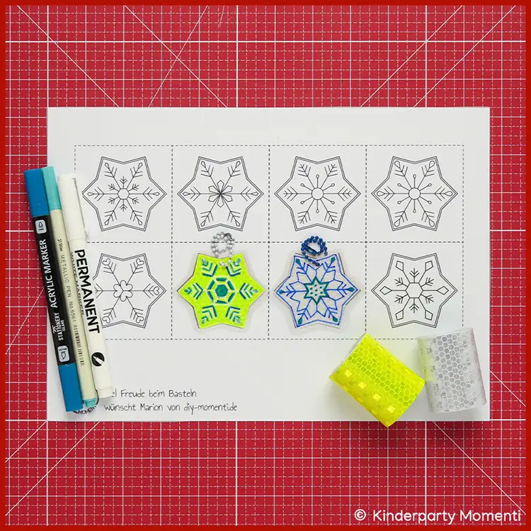 Reflektoren Schneeflocken Anhänger selber machen Bastelvorlage mit Schneeflockenmotiven, zwei bemalte Reflektor Schneeflocken Anhänger in Gelb und Blau, zwei Marker und zwei Rollen Glitzerband auf roter Bastelmatte.