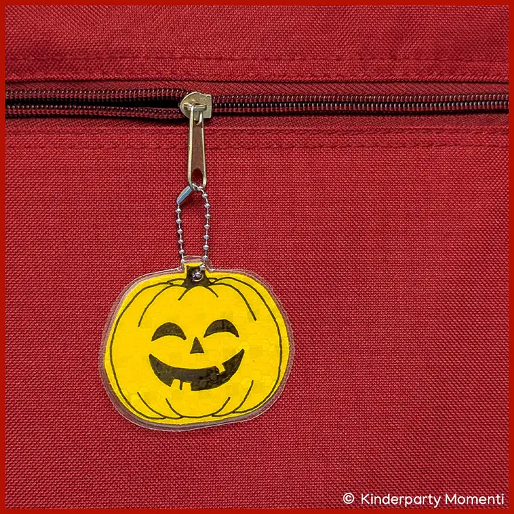 DIY Reflektor Kürbis Anhänger für Taschen Ein gelber Anhänger mit fröhlichem Kürbisgesicht hängt an einer roten Tasche