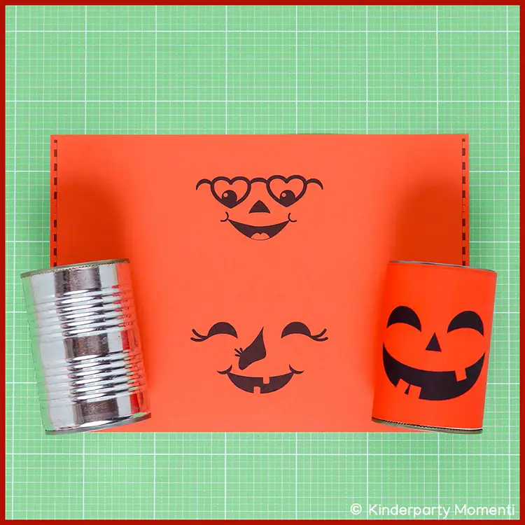 Dosenwerfen aus laminiertem Papier basteln Zwei Dosen, eine blanke und eine mit Kürbis Banderole mit lachendem Gesicht beklebt, neben einer orangefarbenen Vorlage mit zwei aufgemalten lachenden Gesichtern auf einer Bastelmatte